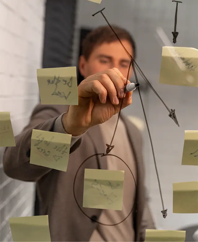 Homem realizando um brainstorming em frente a um quadro de vidro coberto de notas adesivas.